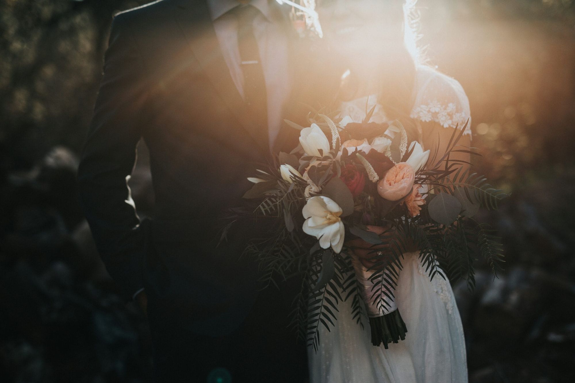 Bouquet de mariée au premier plan avec les mariés en arrière-plan dans la lumière du soleil