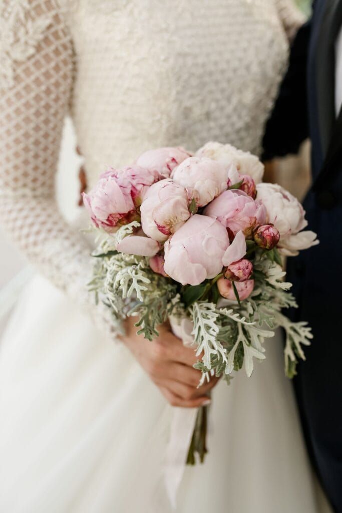 Mariée tenant un bouquet de pivoines rosées avec robe de mariage à manches longues, photographiée dans une ambiance douce et romantique.